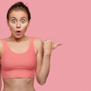 Shocked athletic young woman dressed in pink tanktop, has stupefied expression, points aside with thumb, shows free space for your slogan, has slim muscular body, goes in for sport regularly.